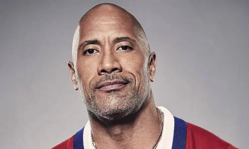 A close-up portrait of a smiling Dwayne "The Rock" Johnson looking directly at the camera against a neutral gray background.