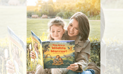Bindi Irwin smiling as she reads her children’s book “You Are a Wildlife Warrior!” with her young daughter outdoors.