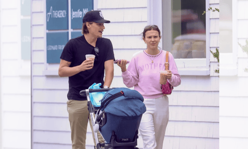 Jake Bongiovi pushes a stroller while Millie Bobby Brown, in a pink “Mother” sweatshirt, walks beside him holding a drink.