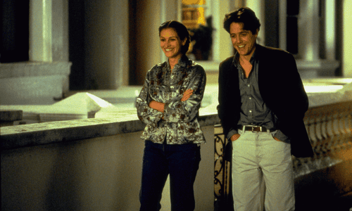 Julia Roberts smiles with crossed arms as Hugh Grant laughs with his hands in his pockets, walking together.