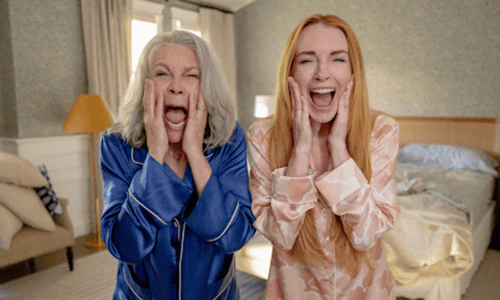 Two women in pajamas holding their faces and laughing in surprise inside a cozy bedroom.