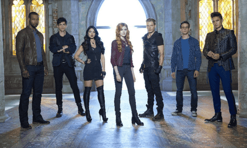 Seven characters from Shadowhunters pose in a gothic hall with large arched windows and stone pillars.