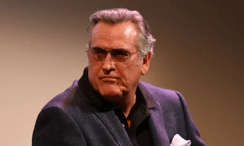  A close-up portrait of actor Bruce Campbell looking sharp in a textured blue blazer and tinted aviator glasses.