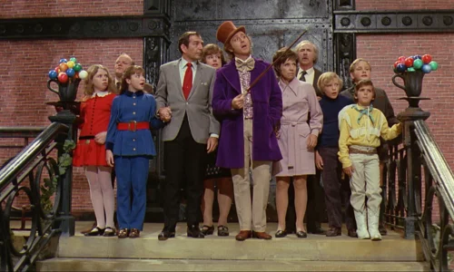 A group of children and Willy Wonka (Gene Wilder) in a purple coat and top hat, stand on a staircase inside a factory set.