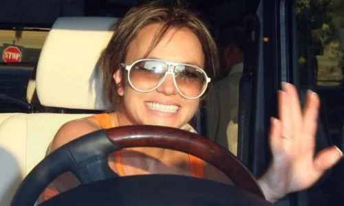 A smiling Britney Spears wearing white sunglasses and waving from the driver's seat of a car.
