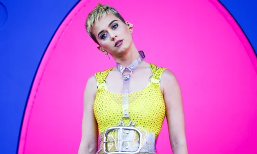 Katy Perry with a short blonde pixie cut wearing a studded yellow dress with a clear plastic harness against a pink and blue background.
