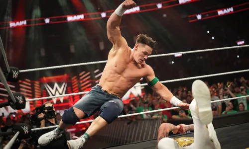 John Cena performing his signature Five Knuckle Shuffle fist drop on an opponent during a WWE RAW wrestling match in a packed arena.