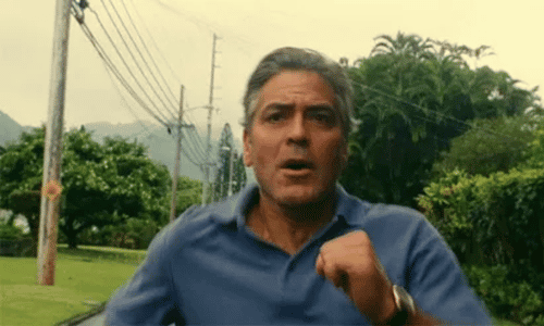 George Clooney running down a Hawaiian street in a patterned shirt in a scene from The Descendants.