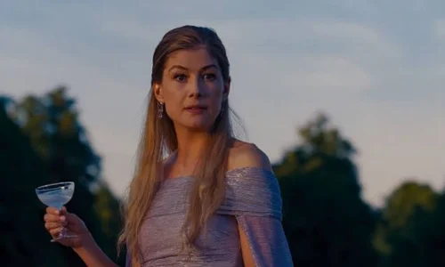 Rosamund Pike in an elegant off-shoulder gown holding a cocktail glass in an outdoor evening scene