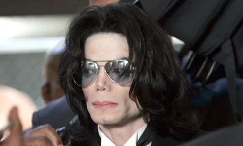 Male singer wearing sunglasses and a dark suit with hands clasped together at a public event