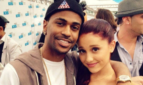 Rapper Big Sean with his arm around singer Ariana Grande as they smile together at an outdoor red carpet event.