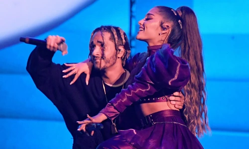 Singer Ariana Grande in a metallic purple outfit performing on a blue-lit stage alongside a male performer.