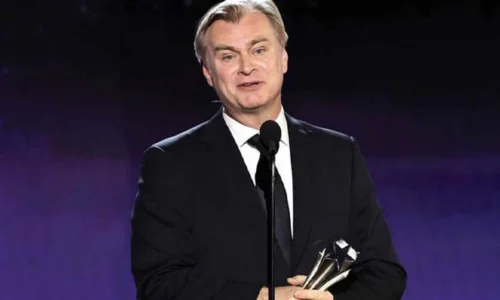 Director Christopher Nolan holds a Critics Choice Award in a black suit and tie against a purple backdrop.
