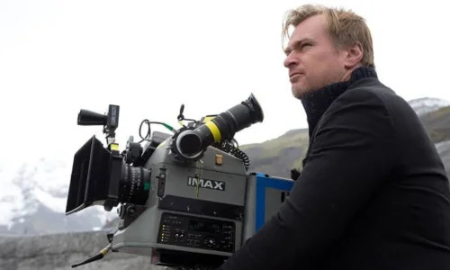 Director Christopher Nolan in profile next to a large IMAX film camera on an outdoor movie set with mountains behind him.