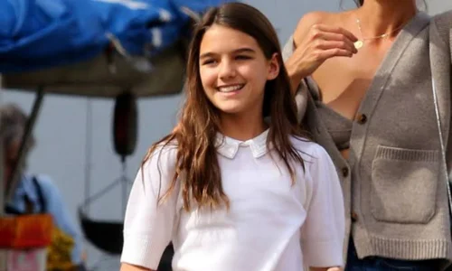 A smiling young Suri Cruise walks outdoors wearing a white short-sleeved polo shirt with a Peter Pan collar.