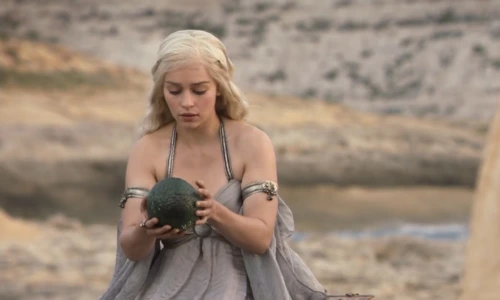 Daenerys Targaryen holding a green dragon egg over a wooden chest.