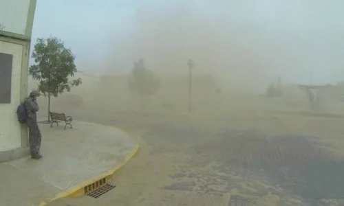 A person with a backpack braces against a building during a thick, brown dust storm obscuring trees and benches in Interstellar.
