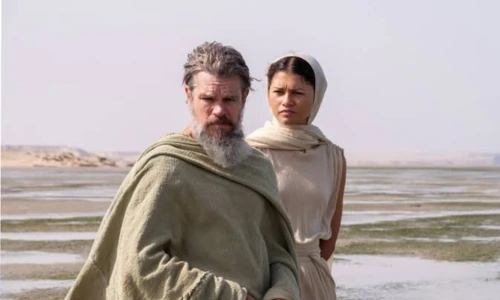 Matt Damon in an olive-green tunic standing next to Zendaya in a cream draped dress against a coastal landscape. 