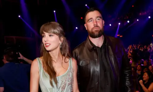 Taylor Swift and Travis Kelce standing together in a dimly lit venue with purple neon lights.
