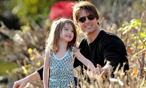 Tom Cruise in sunglasses and a black sweater smiles on the grass next to his young daughter, Suri, in a patterned dress.