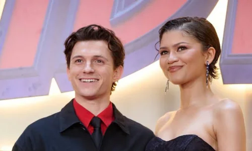 Tom Holland in a black jacket and red shirt stands smiling next to Zendaya in a strapless dress on a red carpet.