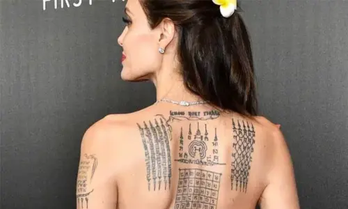 Angelina Jolie looks over her shoulder in profile, showing Sak Yant and text tattoos across her upper back.