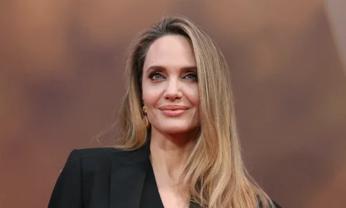 Close-up portrait of Angelina Jolie smiling subtly on a red carpet, wearing a tailored black blazer with her long blonde-highlighted hair draped over her left shoulder.