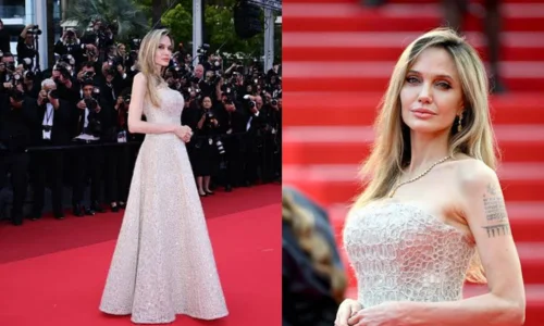 Angelina Jolie stands on a red-carpeted staircase in a strapless light-colored lace gown with intricate sequined patterns.