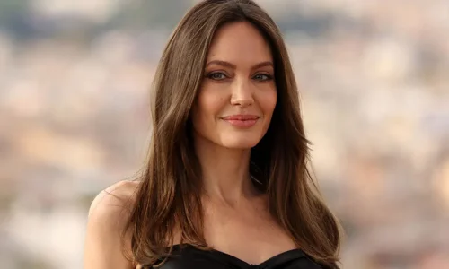 Angelina Jolie smiles subtly with her long brown hair down, wearing a black strapless top outdoors.