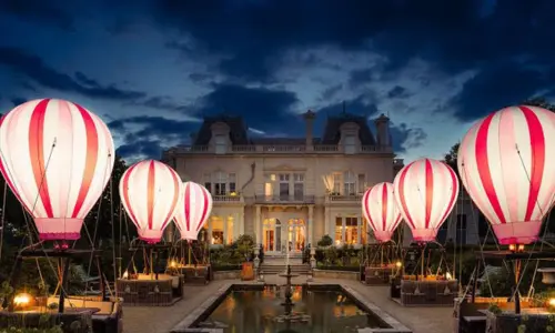 Illuminated pink and white striped hot air balloon dining baskets surround a pool in front of Beaverbrook estate.