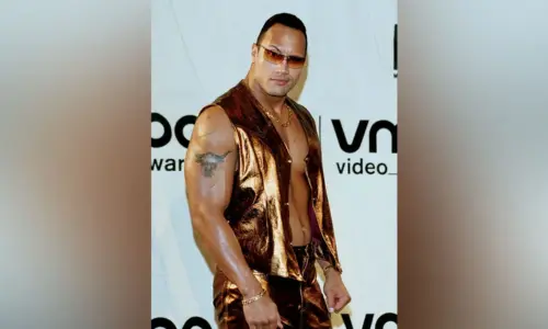Dwayne "The Rock" Johnson poses at an awards show wearing a metallic gold leather vest and sunglasses.