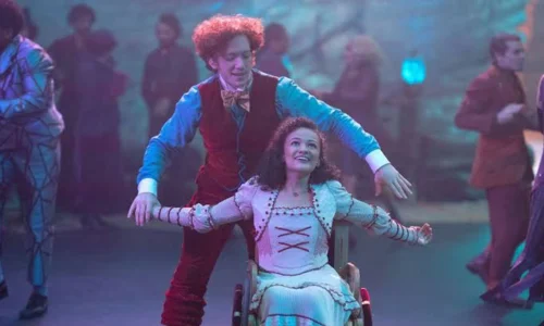 Ethan Slater as Boq standing behind a seated character in a Wicked scene