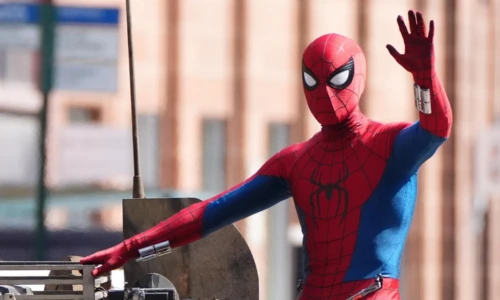 Spider-Man waves with one hand while standing outdoors in his red and blue superhero suit.