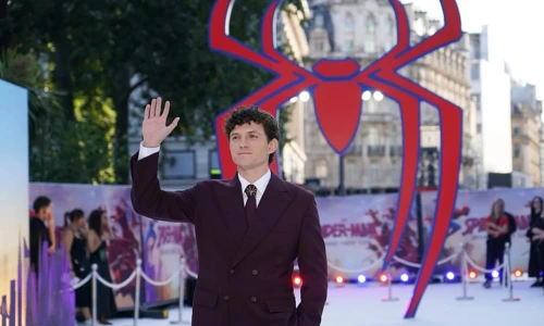 Tom Holland waves on a red carpet in front of a giant Spider-Man logo at the Brand New Day movie premiere.