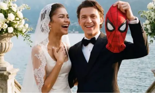 Zendaya laughs in a wedding dress next to Tom Holland, who wears a tuxedo and holds up a Spider-Man mask.