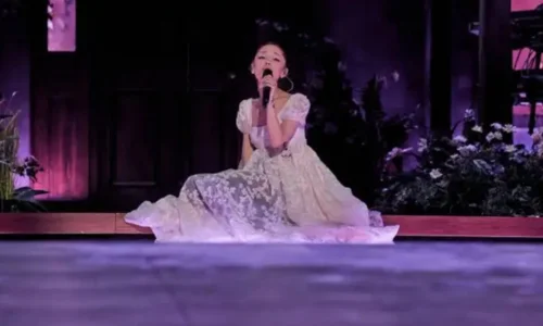 Ariana Grande performing on stage while sitting down, wearing a long, elegant white lace dress with puff sleeves.