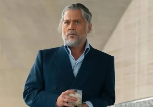 Johnny Depp with grey hair and beard, wearing a navy blue suit and holding a drink glass.