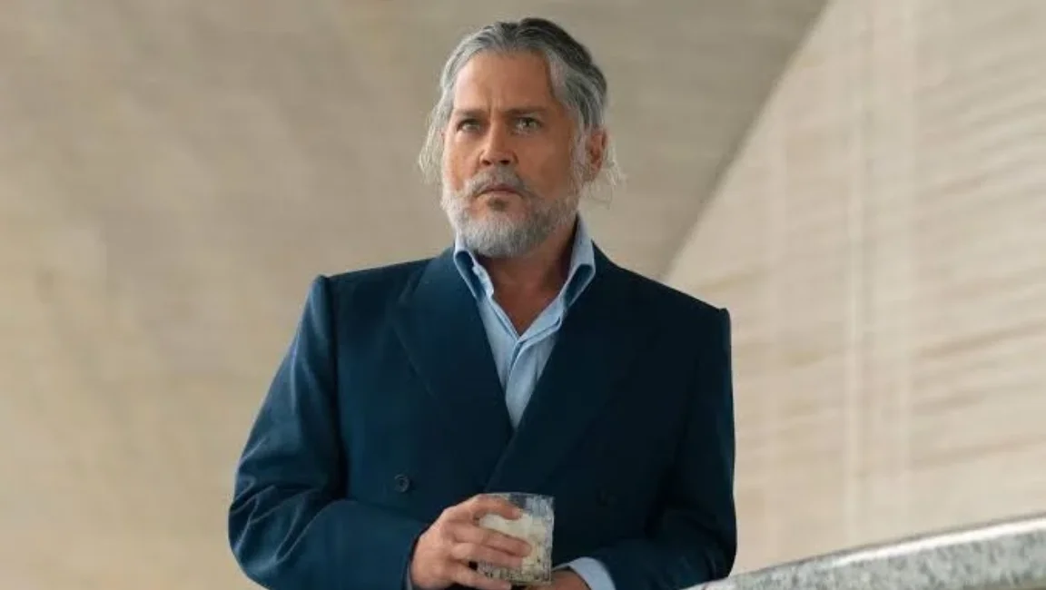 Johnny Depp with grey hair and beard, wearing a navy blue suit and holding a drink glass.