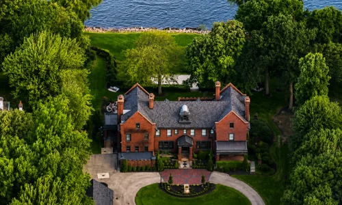 Aerial view of a historic red brick mansion surrounded by trees, a circular driveway, and a blue lake.