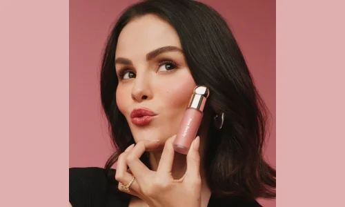 Selena Gomez holding a pink Rare Beauty Soft Pinch Liquid Blush bottle next to her cheek against a pink background.
