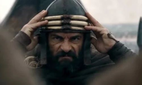 Bearded warrior wearing a dark metal helmet and holding it with both hands while staring intensely ahead.