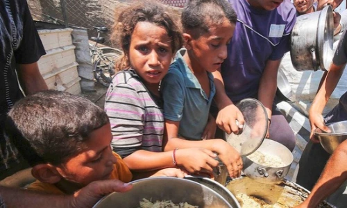 Distressed children hold up empty metal bowls at a crowded food distribution area in a makeshift camp setting.