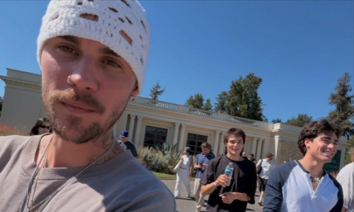 Justin Bieber wears a white knit beanie and chains in an outdoor selfie with friends in front of a white building.