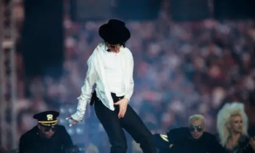 Michael Jackson performs on stage in a white shirt and black fedora surrounded by backup dancers.