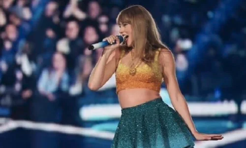 Singer taylor swift performing on stage with a microphone while wearing a gold top and teal skirt